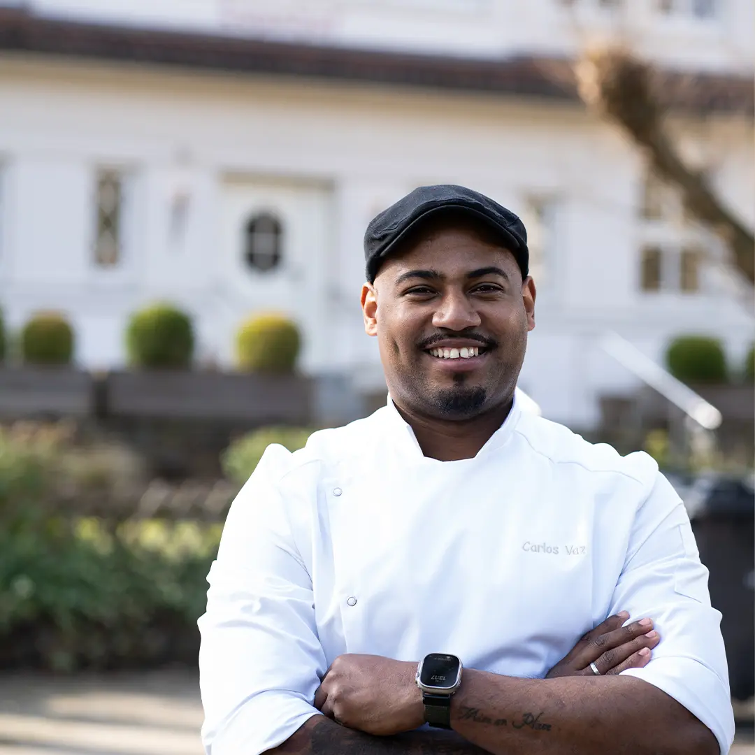 Carlos Vaz mit weißer Kochjacke und schwarzer Schirmmütze lächelnd vorm Lopshof in Dötlingen,