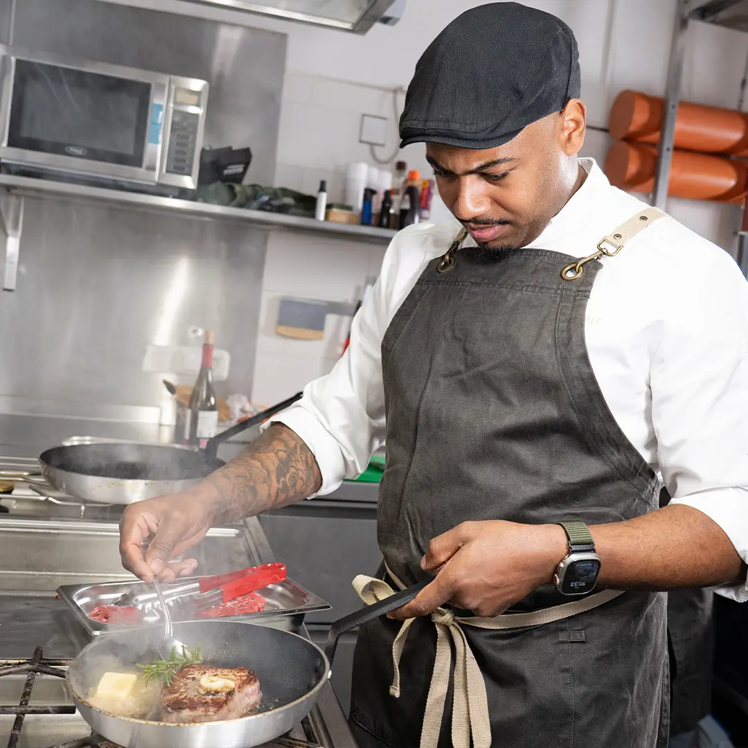 Koch Carlos Vaz in Schürze und Schiebermütze brät ein Steak in einer Pfanne, gibt Butter und Kräuter dazu | Lopshof by Carlos