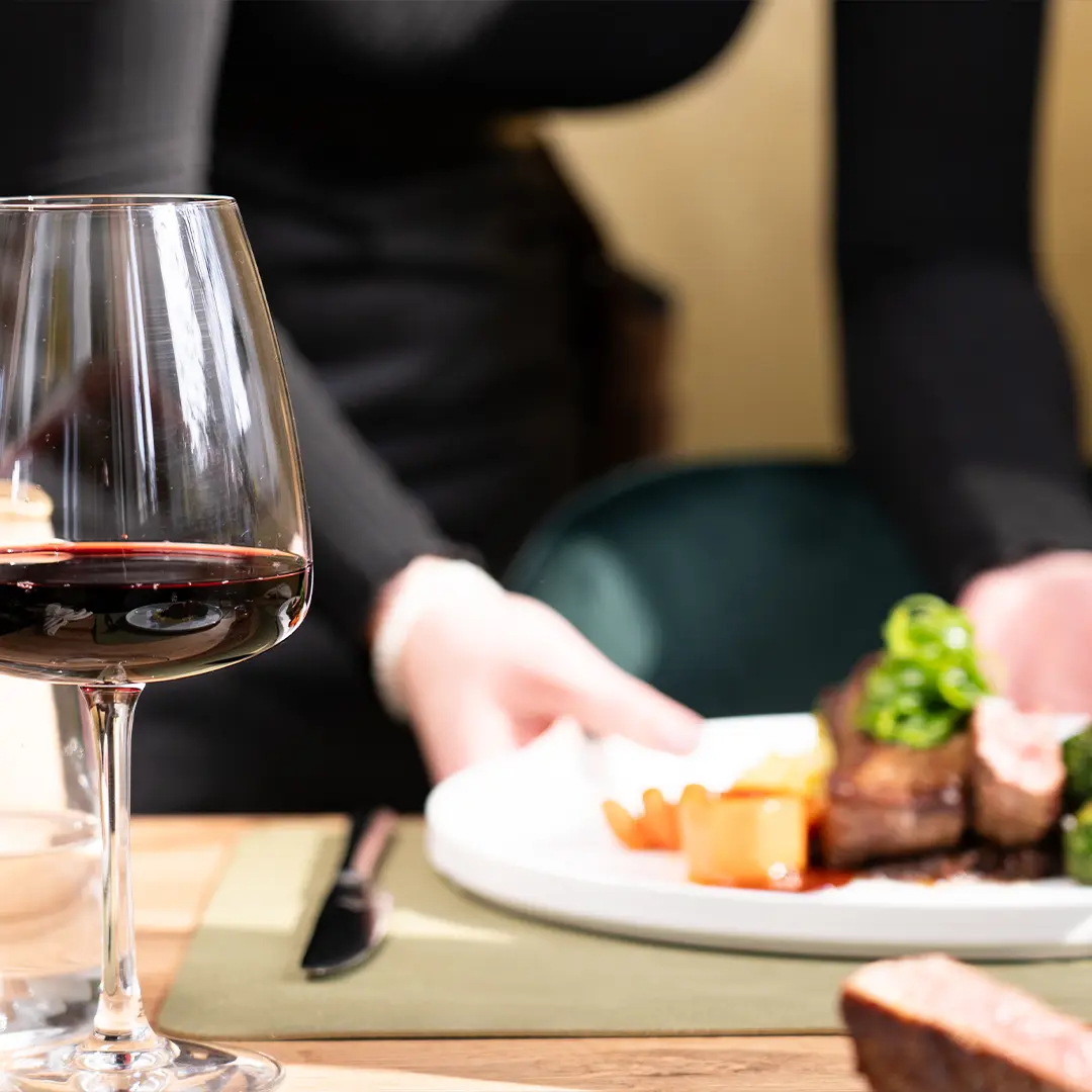 Weinglas mit Rotwein im Vordergrund, im Hintergrund eine Servierperson, die einen Teller mit Fleisch und Gemüse auf den Tisch stellt.