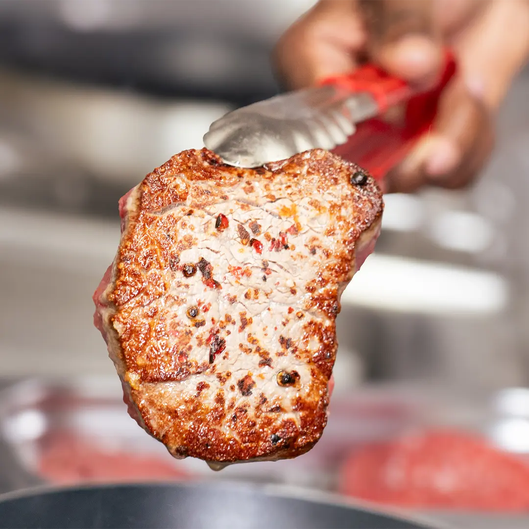 Gegrilltes Steakstück mit würziger Kruste, gehalten von roten Grillzangen über einer Pfanne im Lopshof by Carlos
