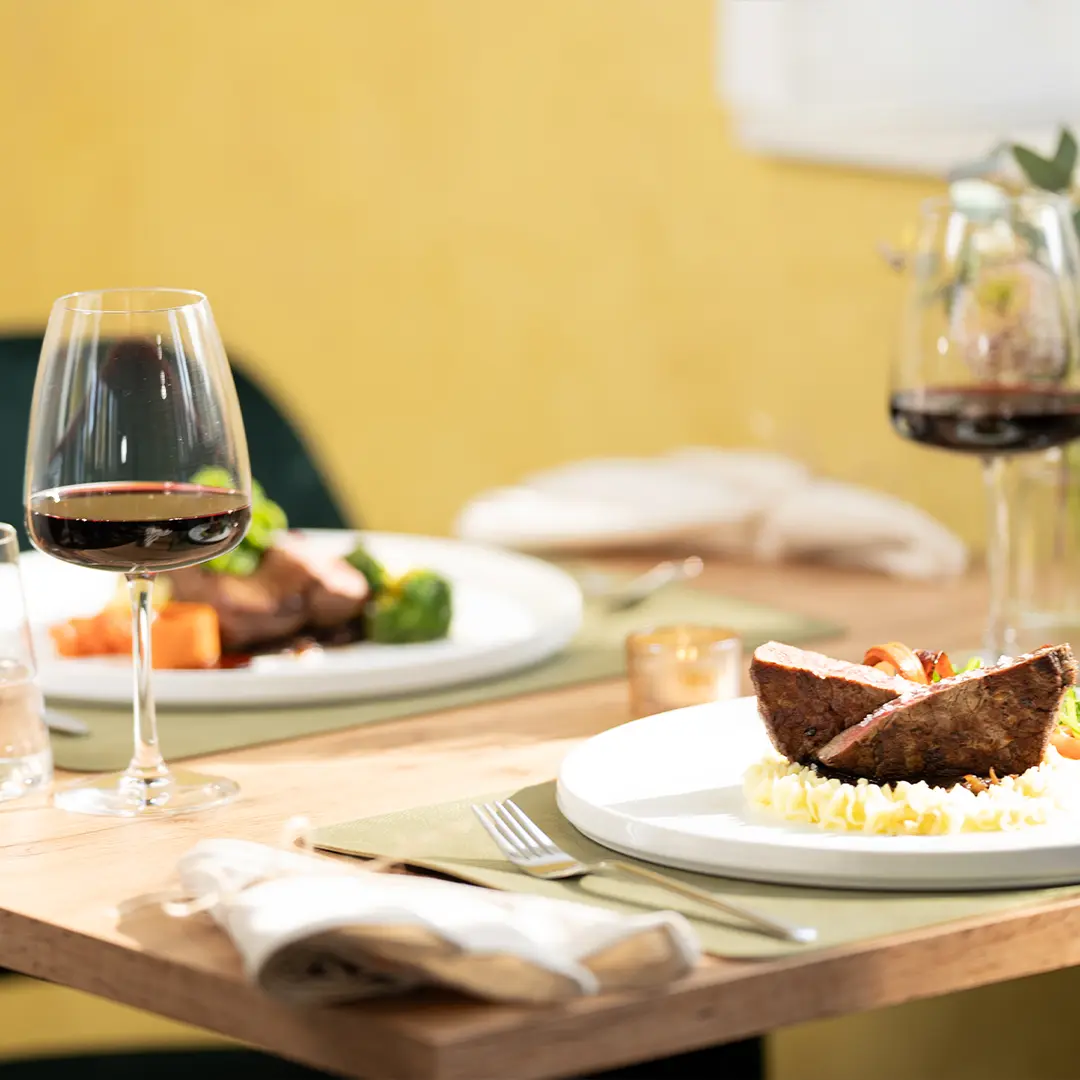 Zwei elegant angerichtete Fleischportionen mit Beilagen auf weißen Tellern und zwei Gläser Rotwein auf einem Tisch im Restaurant Lopshof by Carlos