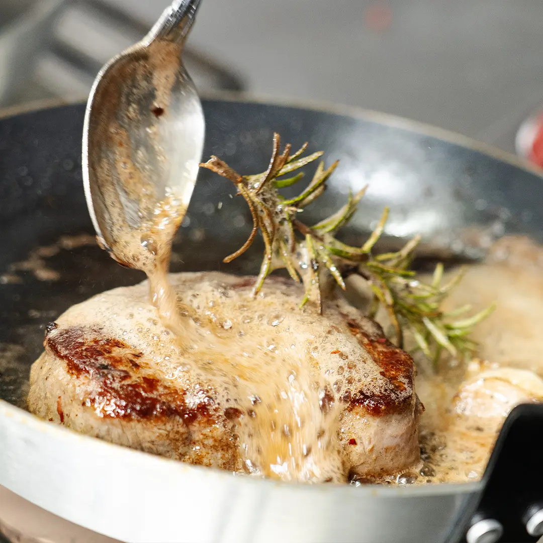 Saftig gebratenes Filetmedaillon in Pfanne, übergossen mit schäumender Bratensauce und mit einem Zweig Rosmarin garniert.