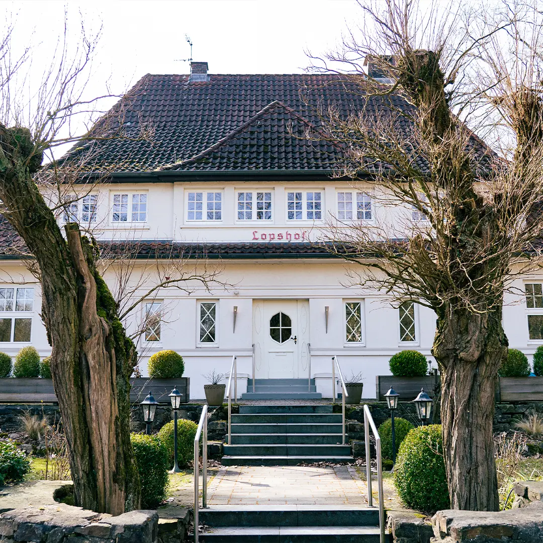 Eingangsfront des Lopshofes, eines weißen Landhauses mit Treppe und Handläufen, zwei knotigen Baumstämmen im Vordergrund