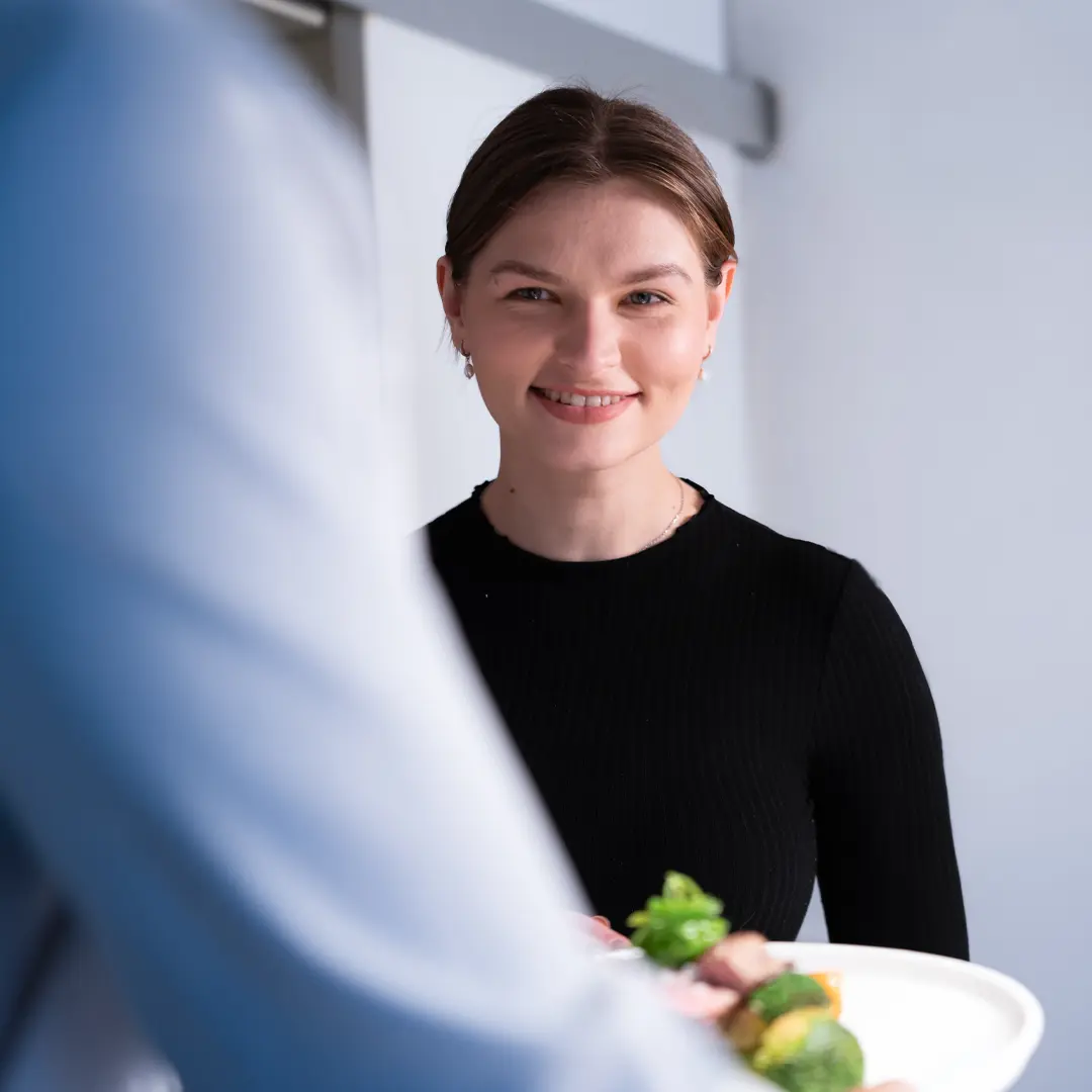 Natascha, Kellnerin im Vordergrund reicht Teller mit grünen Beilagen.