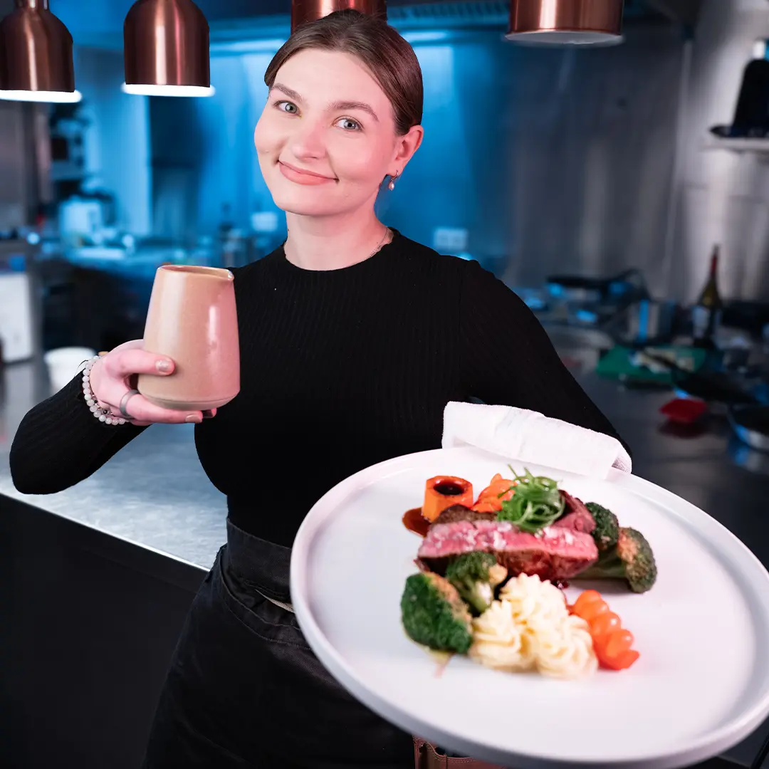 Natascha im Restaurant mit einem Teller fein angerichteten Fleisches, Gemüse und Kartoffelpüree und einer Kaffeetasse in der Hand.