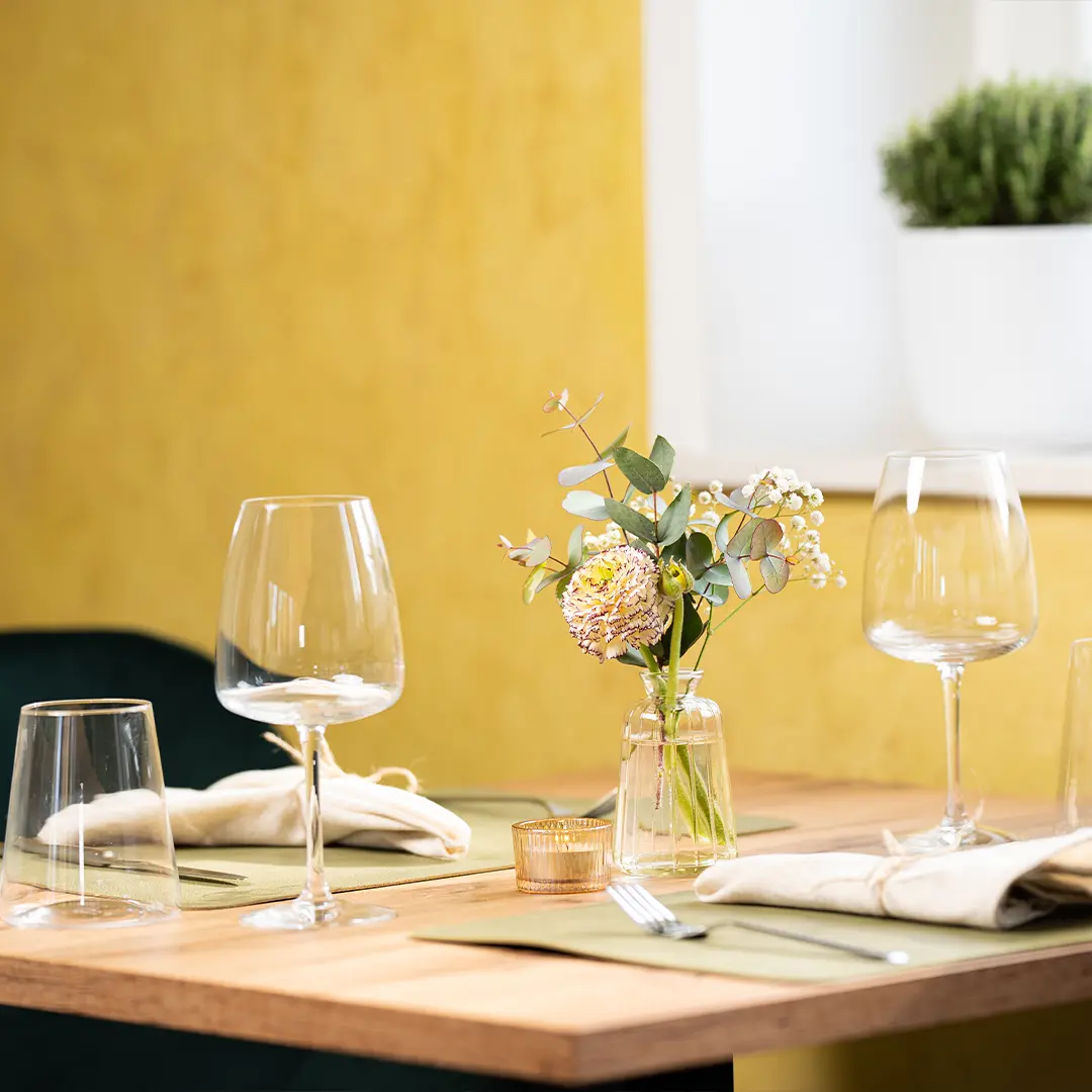 Tisch in hellem Restaurant mit Glasgeschirr, Stoffservietten, kleinem Blumenarrangement in Glasvase und gelber Wand im Hintergrund